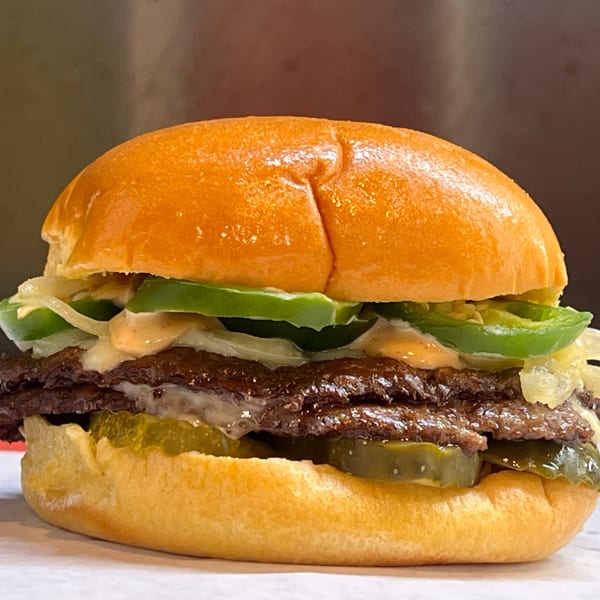 Close-up of a WattaBun burger with two patties, pickles, onions, jalapeños, and sauce.